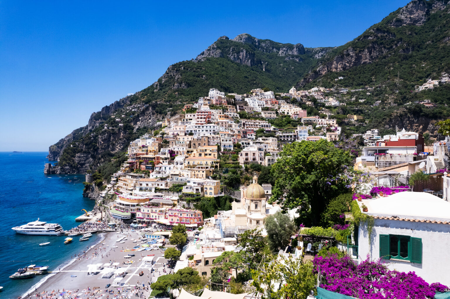Scenic view of Positano, Italy, showcasing colorful cliffside houses, sandy beach, and turquoise waters under a clear blue sky, surrounded by lush greenery and mountains. | Only on SopranoVillas
