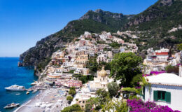 Scenic view of Positano, Italy, showcasing colorful cliffside houses, sandy beach, and turquoise waters under a clear blue sky, surrounded by lush greenery and mountains.
