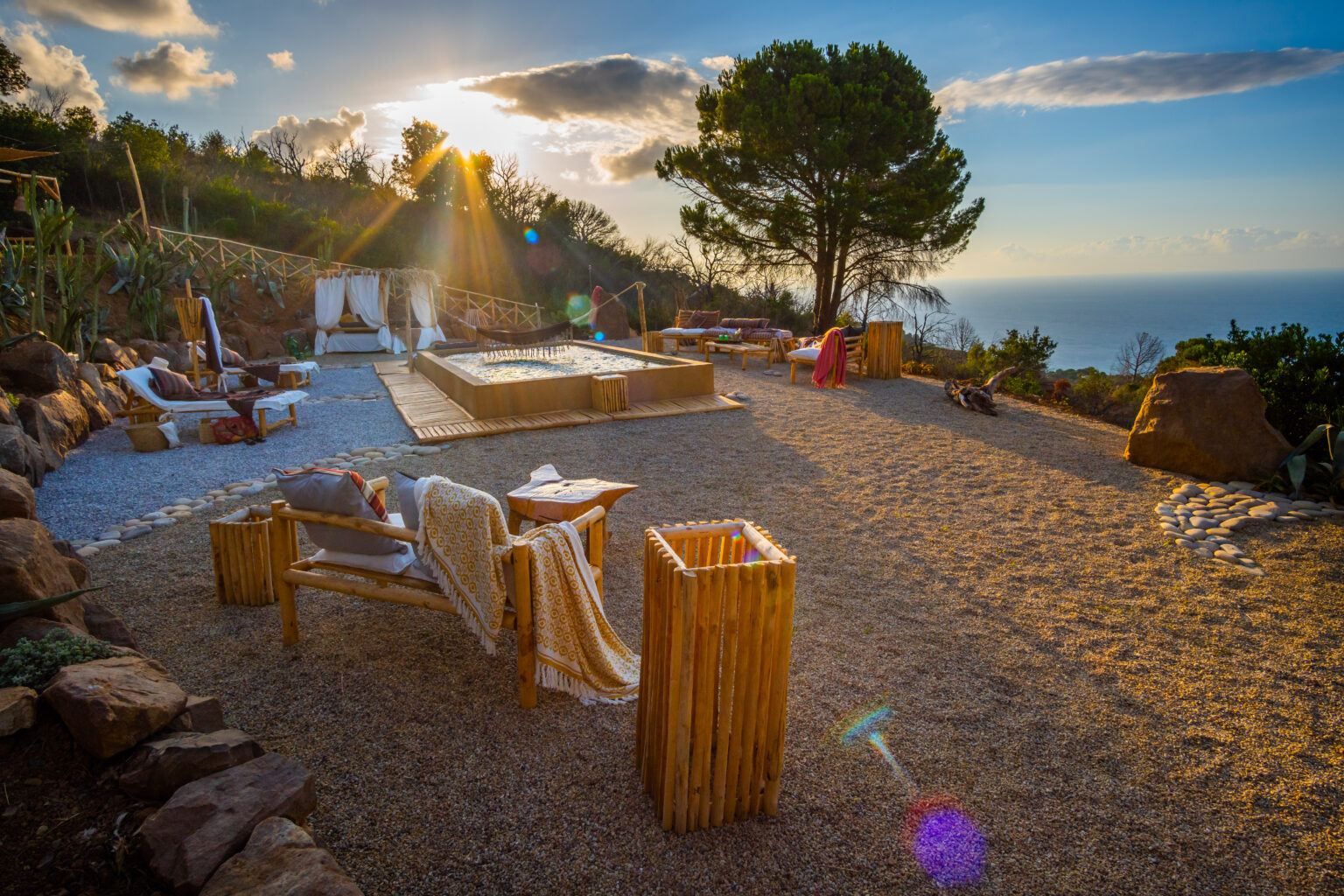 Scenic outdoor relaxation area with wooden furniture, a hot tub, and gravel landscaping, overlooking the ocean at sunset, surrounded by greenery and natural rock features. | Only on SopranoVillas