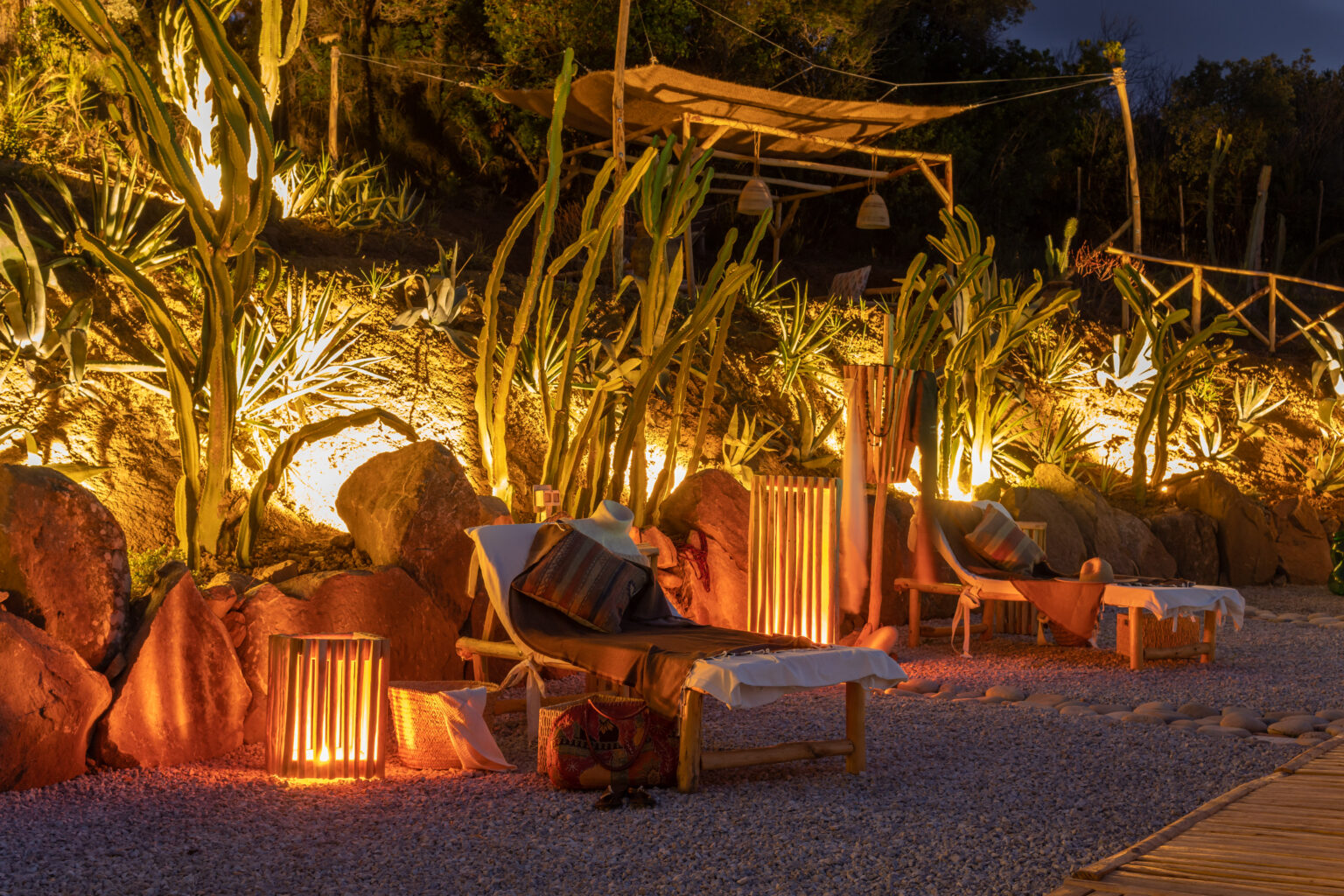 Cozy outdoor lounge area featuring wooden deck chairs, illuminated by warm lighting, surrounded by lush cacti and decorative stones at night. Perfect for relaxation and outdoor gatherings. | Only on SopranoVillas