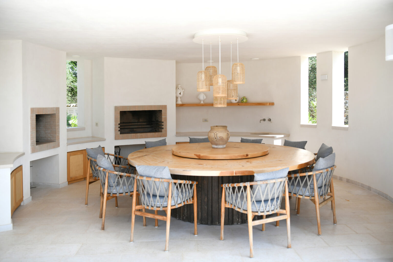 Modern dining area featuring a round wooden table surrounded by stylish wicker chairs. The space includes built-in fireplaces, a minimalist design with white walls, and decorative pendant lighting. Natural light streams through the large windows, enhancing the airy atmosphere. | Only on SopranoVillas