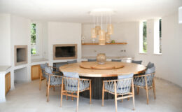 Modern dining area featuring a round wooden table surrounded by stylish wicker chairs. The space includes built-in fireplaces, a minimalist design with white walls, and decorative pendant lighting. Natural light streams through the large windows, enhancing the airy atmosphere.