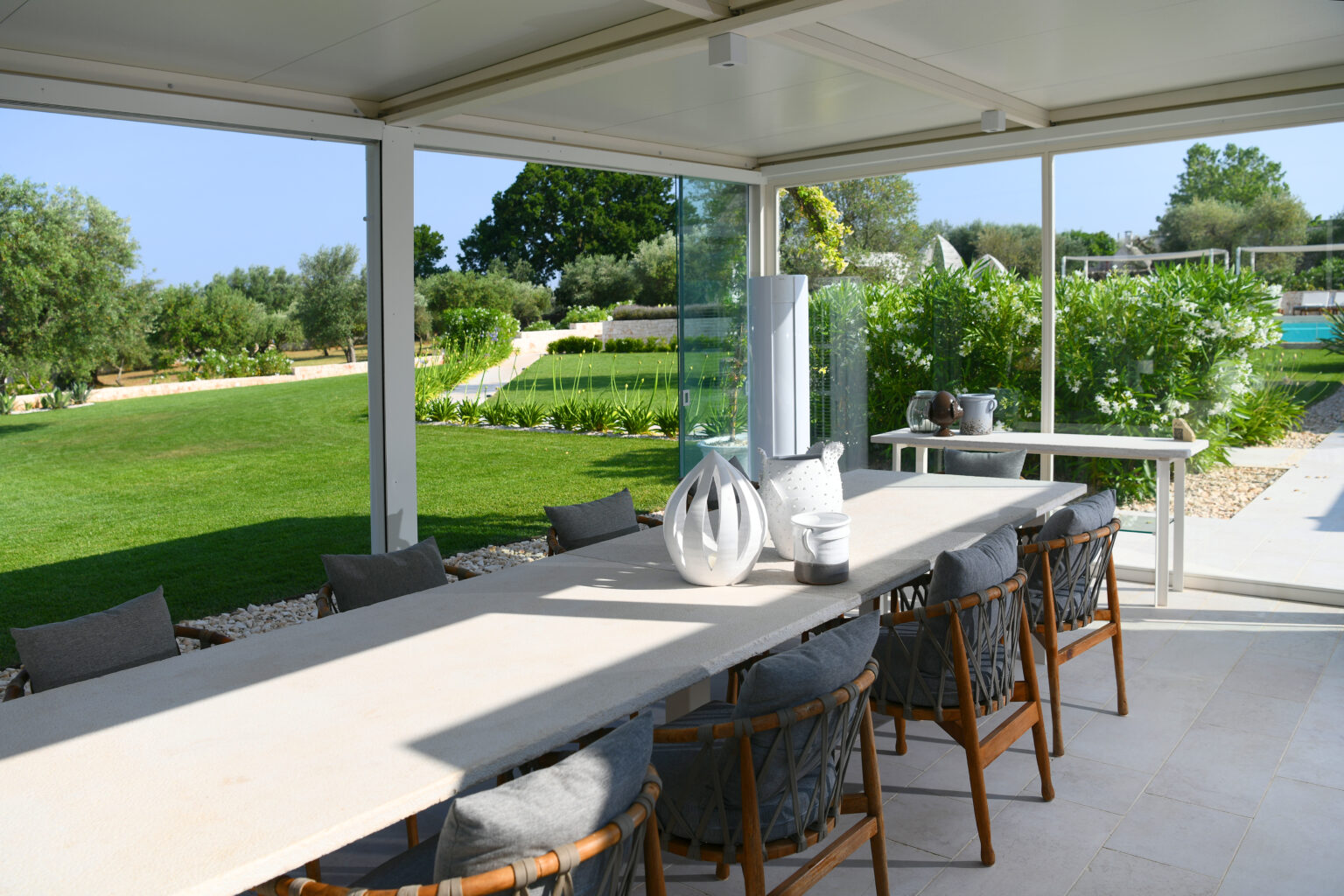 Spacious outdoor dining area featuring a long table with elegant decor, surrounded by comfortable chairs. The space has glass walls offering views of a lush garden and pool area, perfect for alfresco dining. | Only on SopranoVillas