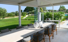 Spacious outdoor dining area featuring a long table with elegant decor, surrounded by comfortable chairs. The space has glass walls offering views of a lush garden and pool area, perfect for alfresco dining.