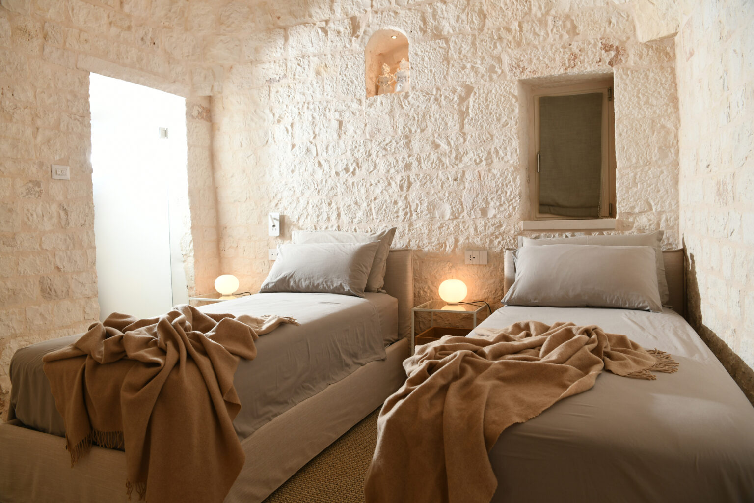 Cozy twin bedroom featuring stone walls, soft gray bedding, and warm lighting. The room is decorated with a neutral-tone blanket draped over the beds, creating a serene and inviting atmosphere. A small window and modern touches, like bedside lamps and wall outlets, enhance the comfort of this stylish space. Perfect for relaxing stays or guest accommodations. | Only on SopranoVillas