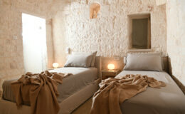 Cozy twin bedroom featuring stone walls, soft gray bedding, and warm lighting. The room is decorated with a neutral-tone blanket draped over the beds, creating a serene and inviting atmosphere. A small window and modern touches, like bedside lamps and wall outlets, enhance the comfort of this stylish space. Perfect for relaxing stays or guest accommodations.