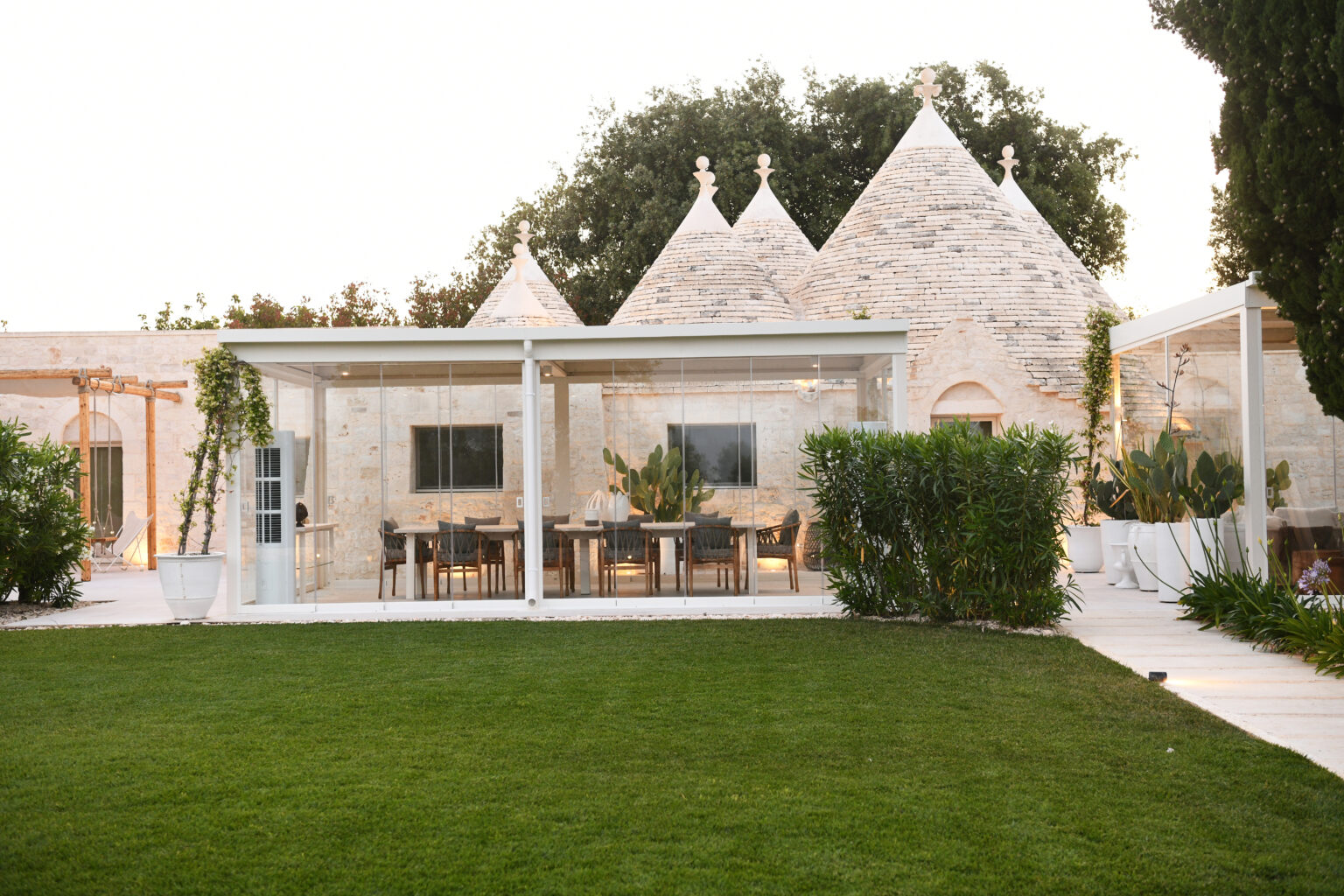 Modern glass-enclosed dining area adjacent to traditional trullo buildings, surrounded by lush green lawn and ornamental plants, showcasing a blend of contemporary and rustic architecture. | Only on SopranoVillas