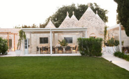 Modern glass-enclosed dining area adjacent to traditional trullo buildings, surrounded by lush green lawn and ornamental plants, showcasing a blend of contemporary and rustic architecture.