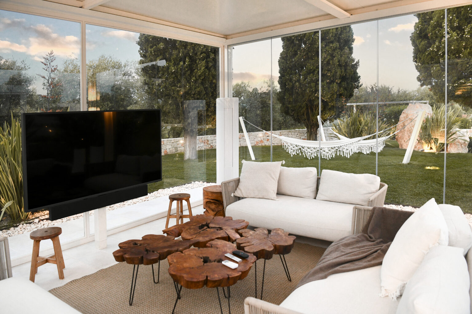 Modern outdoor living space featuring a cozy seating area with a wooden coffee table, a large flat-screen TV, and a view of a green garden with a hammock. Natural light fills the room through glass walls, enhancing the inviting atmosphere. | Only on SopranoVillas
