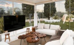 Modern outdoor living space featuring a cozy seating area with a wooden coffee table, a large flat-screen TV, and a view of a green garden with a hammock. Natural light fills the room through glass walls, enhancing the inviting atmosphere.