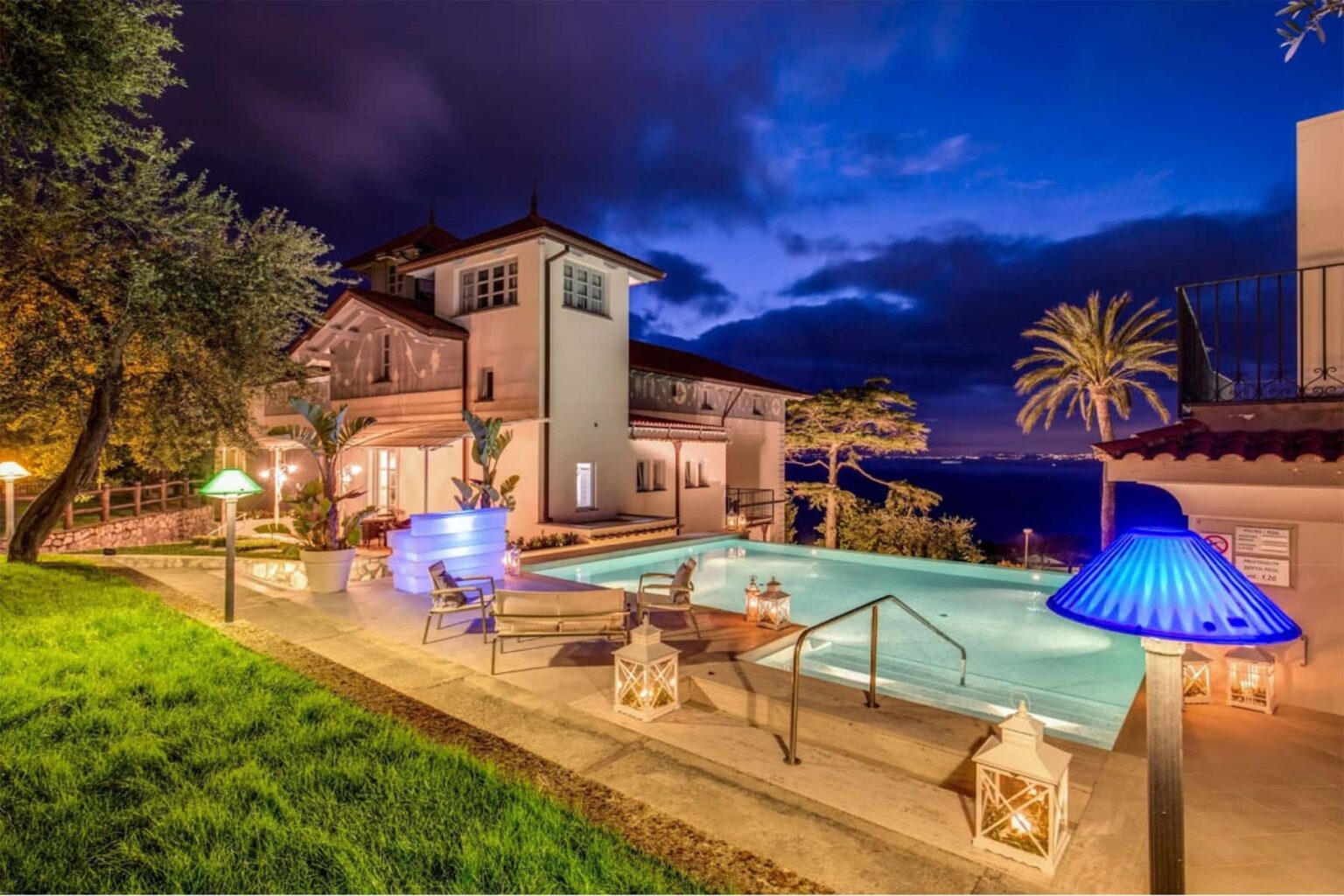 Luxury villa with a stunning pool at dusk, surrounded by lush greenery and palm trees, featuring ambient lighting and a scenic ocean view. Perfect for relaxation and outdoor gatherings. | Only on SopranoVillas