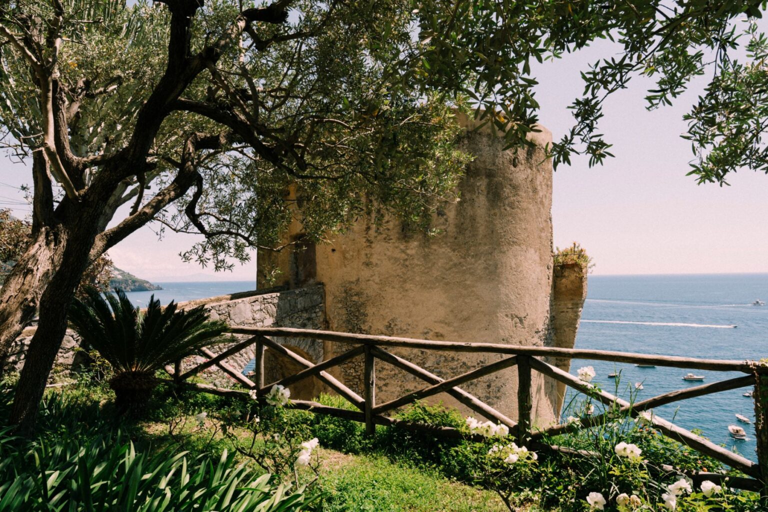 Scenic view of a historic tower by the sea, surrounded by lush greenery and a wooden fence, highlighting luxury villas for rent in Italy's picturesque coastal landscape. | Only on SopranoVillas