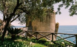 Scenic view of a historic tower by the sea, surrounded by lush greenery and a wooden fence, highlighting luxury villas for rent in Italy's picturesque coastal landscape.