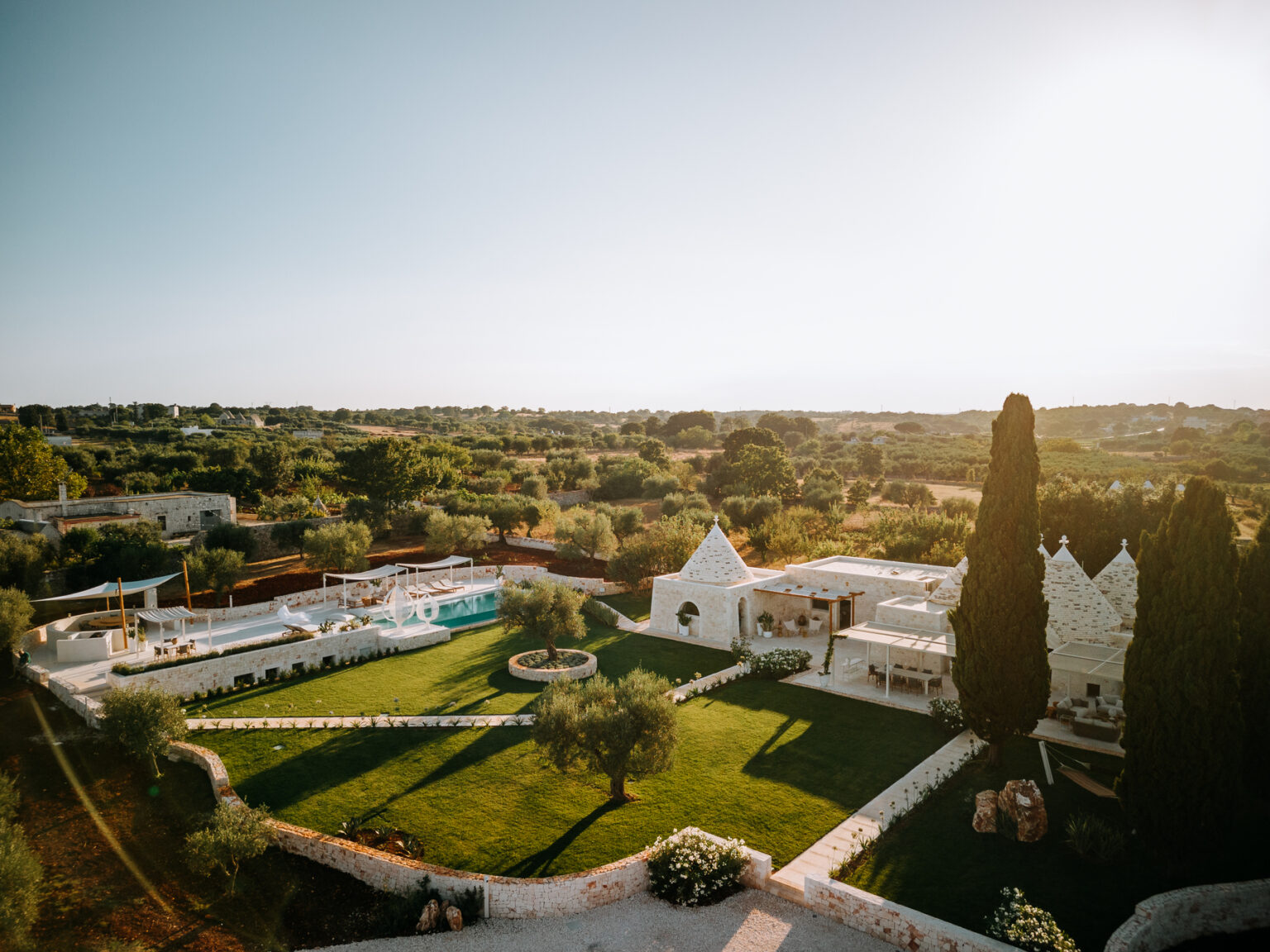 Scenic view of a luxurious villa with a pool and landscaped gardens, surrounded by olive trees and rolling hills under a clear sky. The villa features traditional architecture with unique cone-shaped roofs, ideal for a relaxing getaway in a tranquil setting. | Only on SopranoVillas
