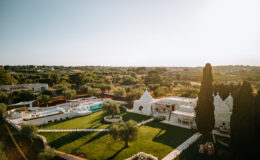 Scenic view of a luxurious villa with a pool and landscaped gardens, surrounded by olive trees and rolling hills under a clear sky. The villa features traditional architecture with unique cone-shaped roofs, ideal for a relaxing getaway in a tranquil setting.