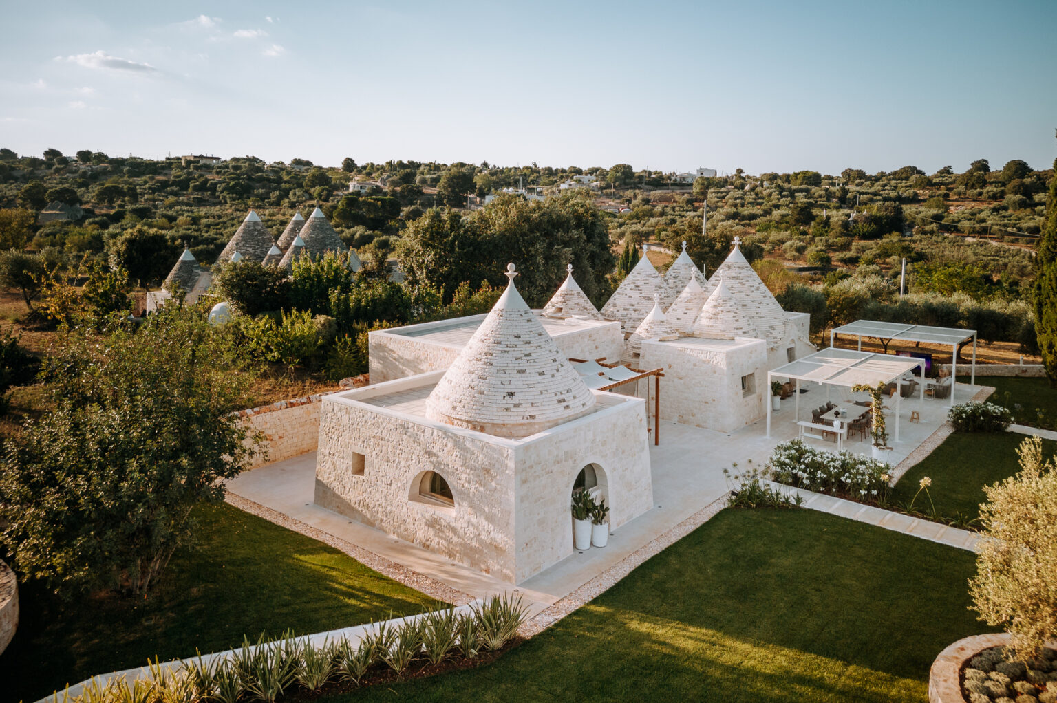 Aerial view of traditional trullo houses with conical roofs surrounded by lush greenery in a rural setting. The image showcases the unique architecture and serene landscape of the Apulia region in Italy. | Only on SopranoVillas