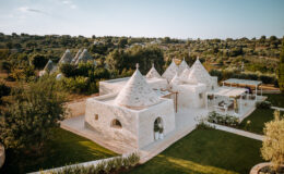 Aerial view of traditional trullo houses with conical roofs surrounded by lush greenery in a rural setting. The image showcases the unique architecture and serene landscape of the Apulia region in Italy.