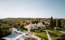 Aerial view of a luxury villa with a pool surrounded by lush greenery and olive trees, featuring traditional trullo architecture in a serene landscape. Ideal for vacation rentals and outdoor gatherings.