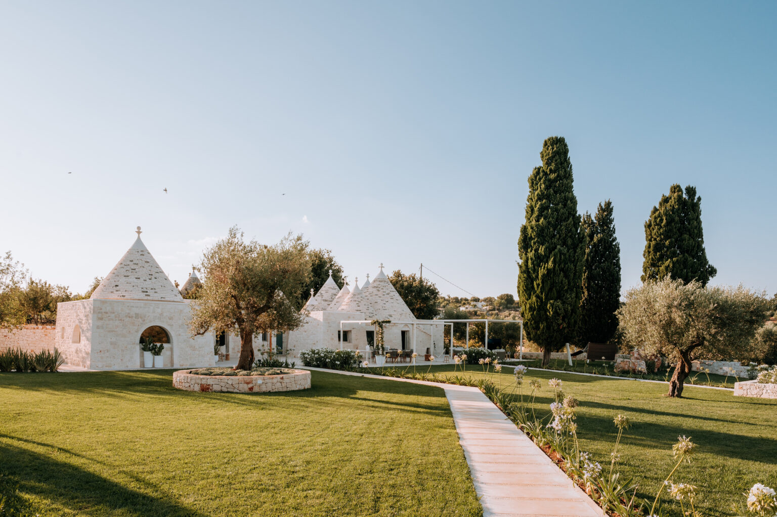 Tranquil white stone villa in a lush green garden, featuring traditional trullo architecture, cypress trees, and a winding pathway under a clear blue sky. | Only on SopranoVillas