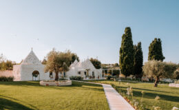 Tranquil white stone villa in a lush green garden, featuring traditional trullo architecture, cypress trees, and a winding pathway under a clear blue sky.