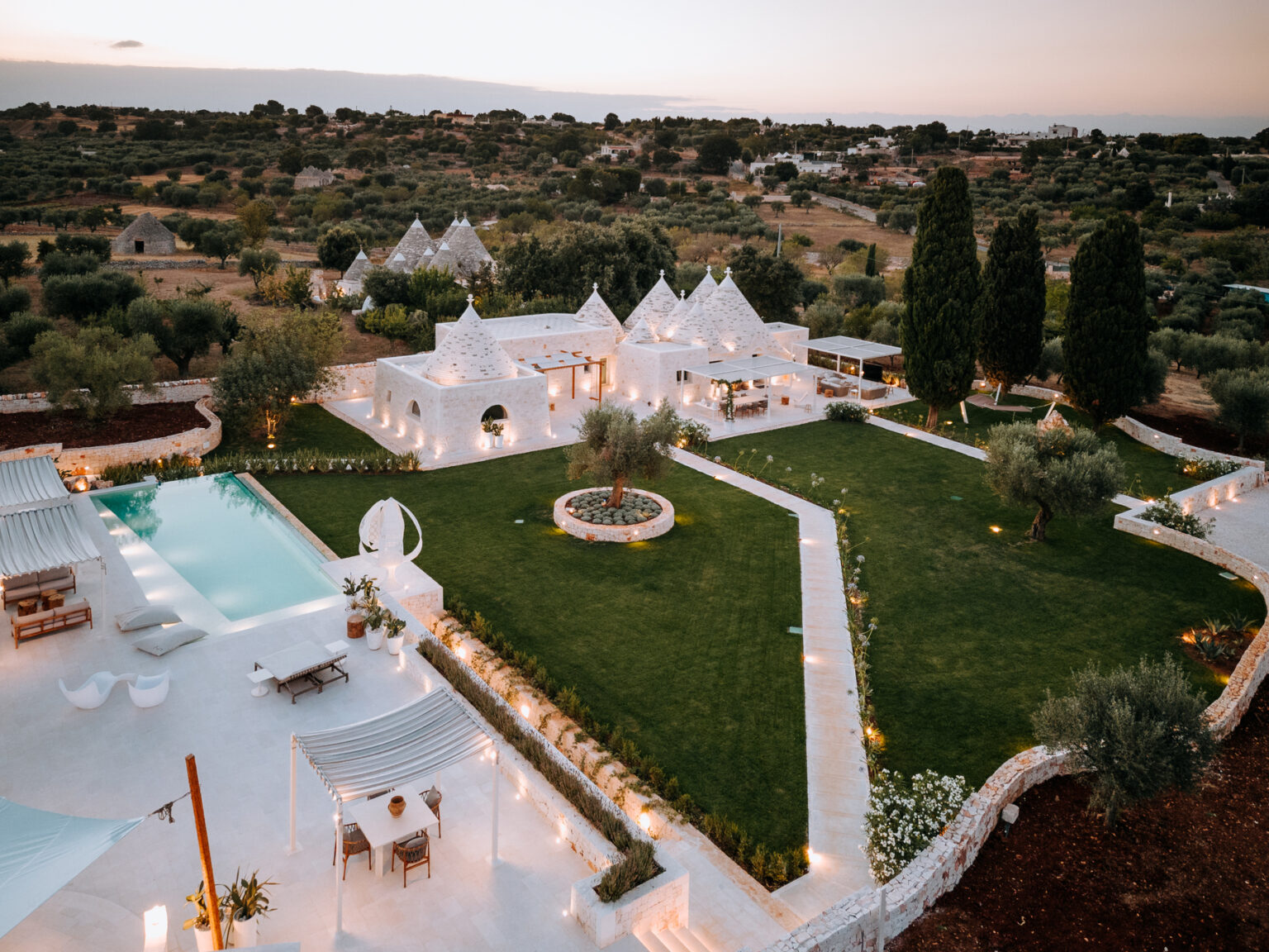 Aerial view of a luxurious white villa surrounded by lush greenery and a well-maintained garden, featuring a sparkling swimming pool and elegant outdoor seating areas, set against a picturesque sunset in a serene landscape. | Only on SopranoVillas