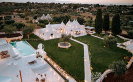 Aerial view of a luxurious white villa surrounded by lush greenery and a well-maintained garden, featuring a sparkling swimming pool and elegant outdoor seating areas, set against a picturesque sunset in a serene landscape.