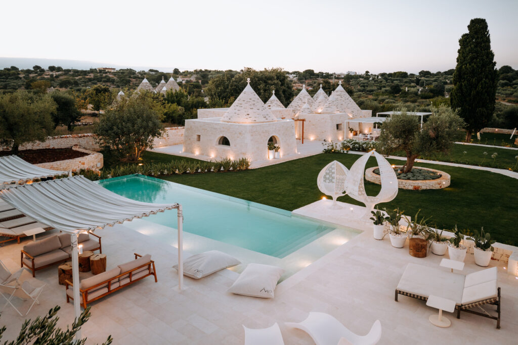 Luxurious white villa with a large swimming pool, surrounded by lush greenery and modern outdoor furniture, set against a serene landscape at sunset. | Only on SopranoVillas