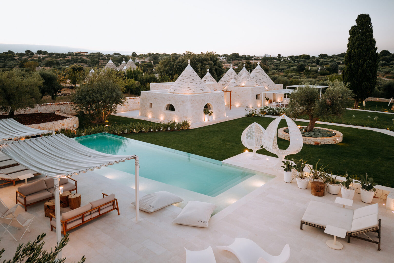 Luxurious white villa with a large swimming pool, surrounded by lush greenery and modern outdoor furniture, set against a serene landscape at sunset. | Only on SopranoVillas