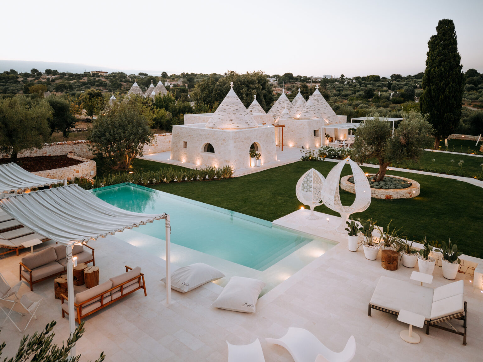 Luxurious villa with a modern pool surrounded by lush greenery, featuring traditional trullo architecture, stylish outdoor seating, and elegant landscaping in a tranquil setting. | Only on SopranoVillas