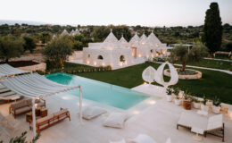 Luxurious white villa with a large swimming pool, surrounded by lush greenery and modern outdoor furniture, set against a serene landscape at sunset.