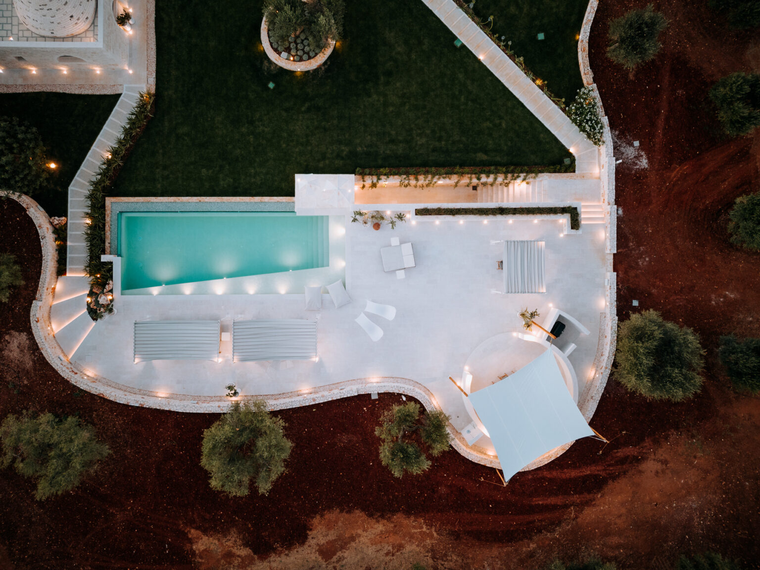 Aerial view of a modern outdoor pool area featuring a turquoise swimming pool, lounge chairs, and a white canopy, surrounded by landscaped greenery and illuminated pathways. | Only on SopranoVillas