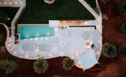 Aerial view of a modern outdoor pool area featuring a turquoise swimming pool, lounge chairs, and a white canopy, surrounded by landscaped greenery and illuminated pathways.