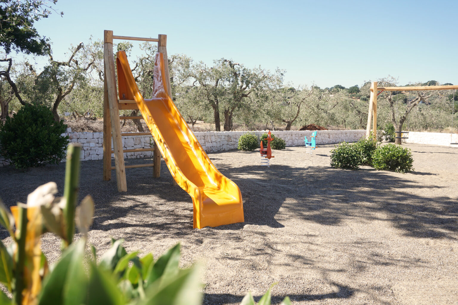 Colorful yellow slide in a sunny outdoor playground surrounded by olive trees and shrubs, featuring sand underfoot and swings in the background. Ideal for children's recreational activities. | Only on SopranoVillas