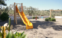 Colorful yellow slide in a sunny outdoor playground surrounded by olive trees and shrubs, featuring sand underfoot and swings in the background. Ideal for children's recreational activities.