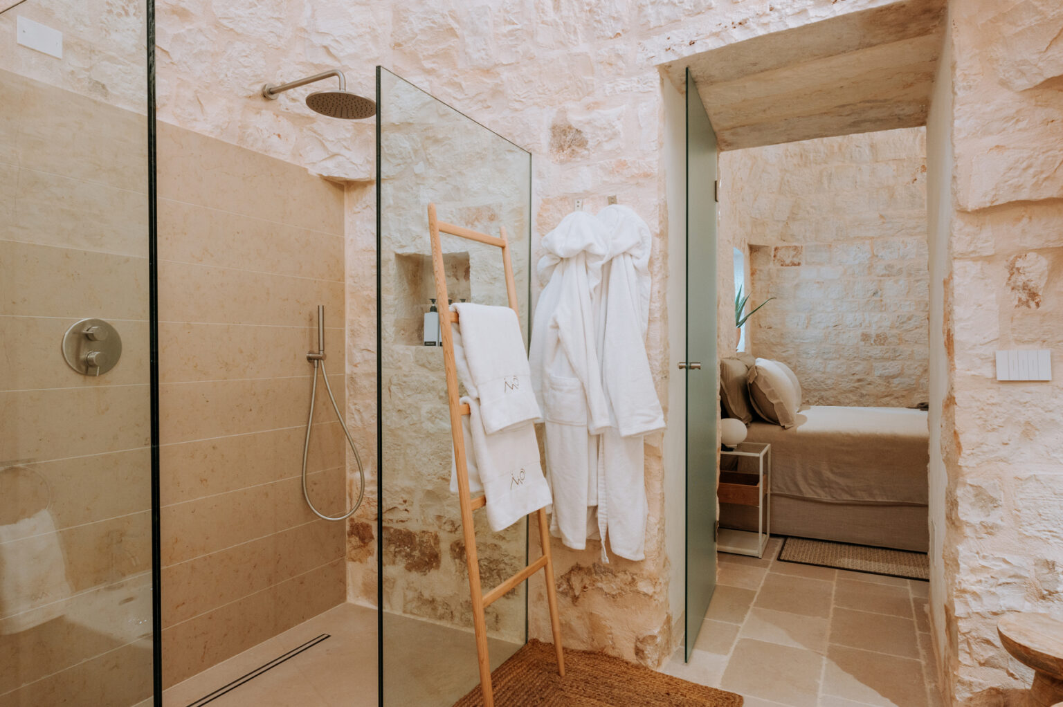 Modern bathroom featuring a glass shower, stone walls, and plush bathrobes hanging on a wooden rack, with a cozy bedroom visible in the background. | Only on SopranoVillas