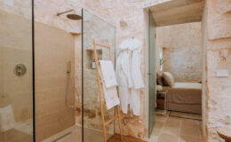 Modern bathroom featuring a glass shower, stone walls, and plush bathrobes hanging on a wooden rack, with a cozy bedroom visible in the background.