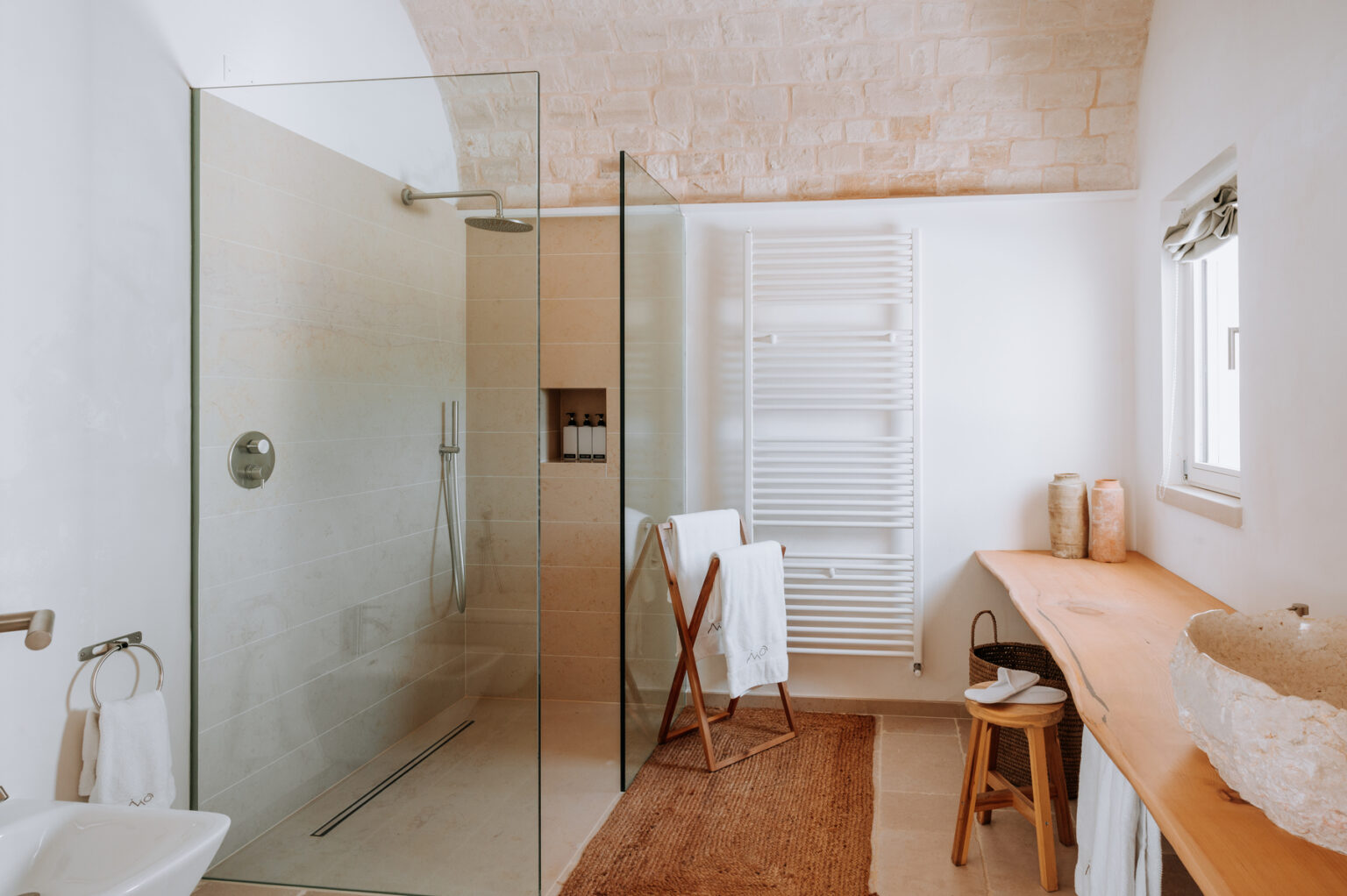 Modern bathroom featuring a glass shower enclosure, wooden countertop, and stylish decor. The spacious design includes a towel rack, a stool, and natural light from a window, creating a serene atmosphere. | Only on SopranoVillas
