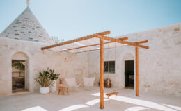 Tranquil outdoor patio featuring a wooden pergola, stone walls, and a cozy seating area, illuminated by bright sunlight, ideal for relaxation and social gatherings.