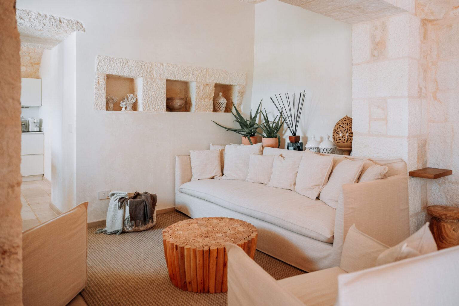 Bright and airy living room featuring a large white sofa, wooden coffee table, and decorative plants. The minimalist decor includes textured walls and natural materials, creating a cozy and inviting atmosphere. | Only on SopranoVillas