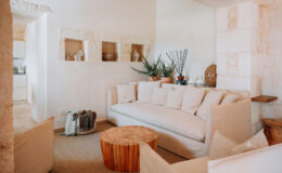 Bright and airy living room featuring a large white sofa, wooden coffee table, and decorative plants. The minimalist decor includes textured walls and natural materials, creating a cozy and inviting atmosphere.