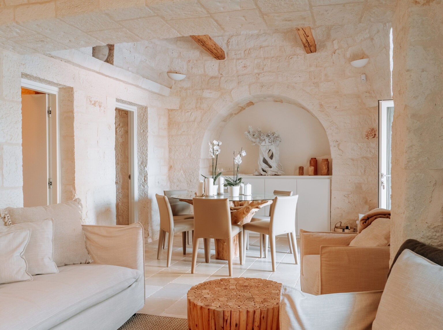 Cozy dining area in a modern stone house featuring a round wooden table, beige chairs, and elegant decor including orchids and candles, complemented by natural light and rustic wooden beams. | Only on SopranoVillas