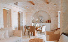 Cozy dining area in a modern stone house featuring a round wooden table, beige chairs, and elegant decor including orchids and candles, complemented by natural light and rustic wooden beams.