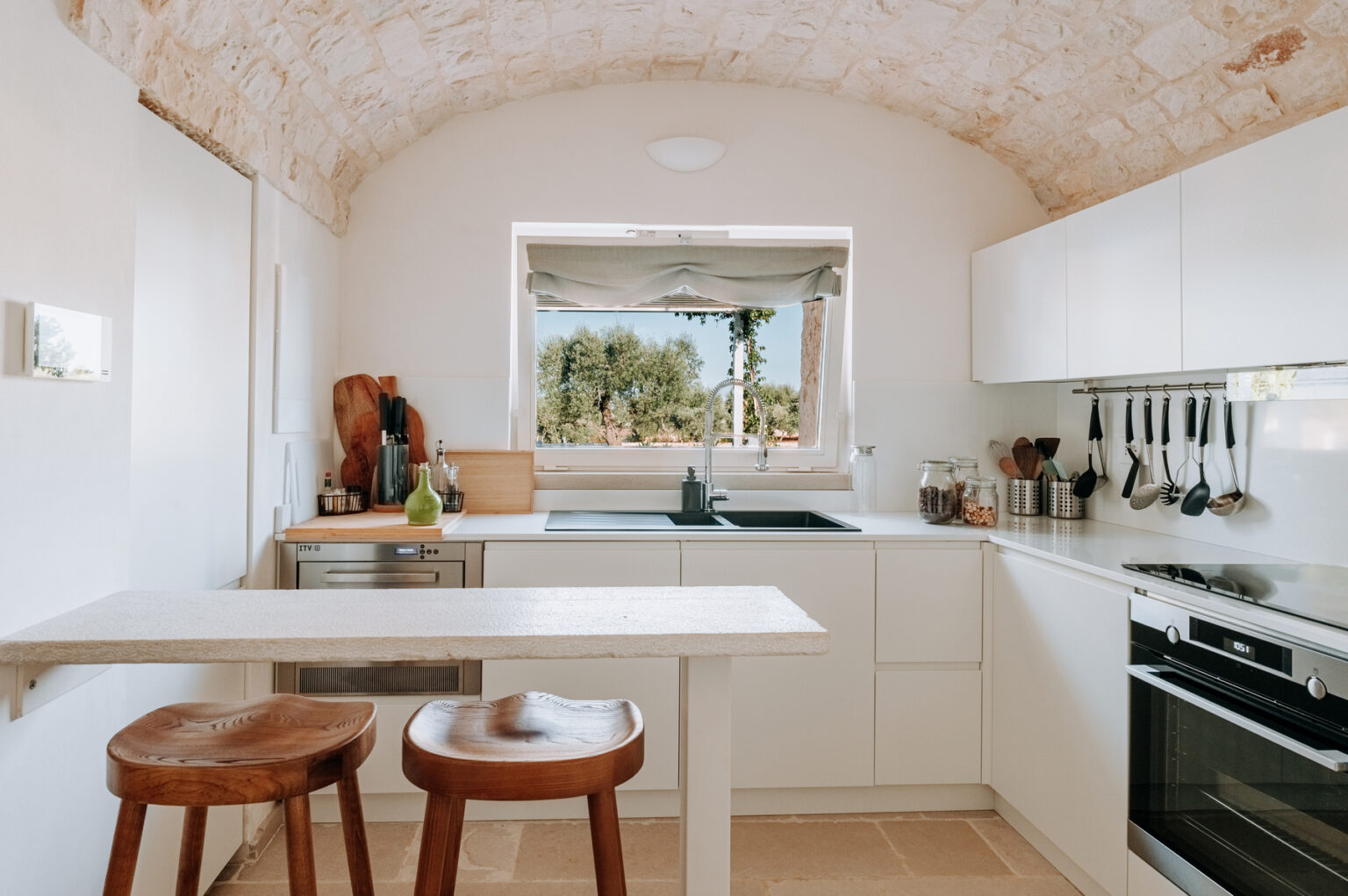 Modern kitchen with white cabinetry, a minimalist design, and an arched ceiling. Features a central island with two wooden stools, large window overlooking greenery, and stainless steel appliances, creating a bright and inviting cooking space. | Only on SopranoVillas