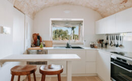 Modern kitchen with white cabinetry, a minimalist design, and an arched ceiling. Features a central island with two wooden stools, large window overlooking greenery, and stainless steel appliances, creating a bright and inviting cooking space.