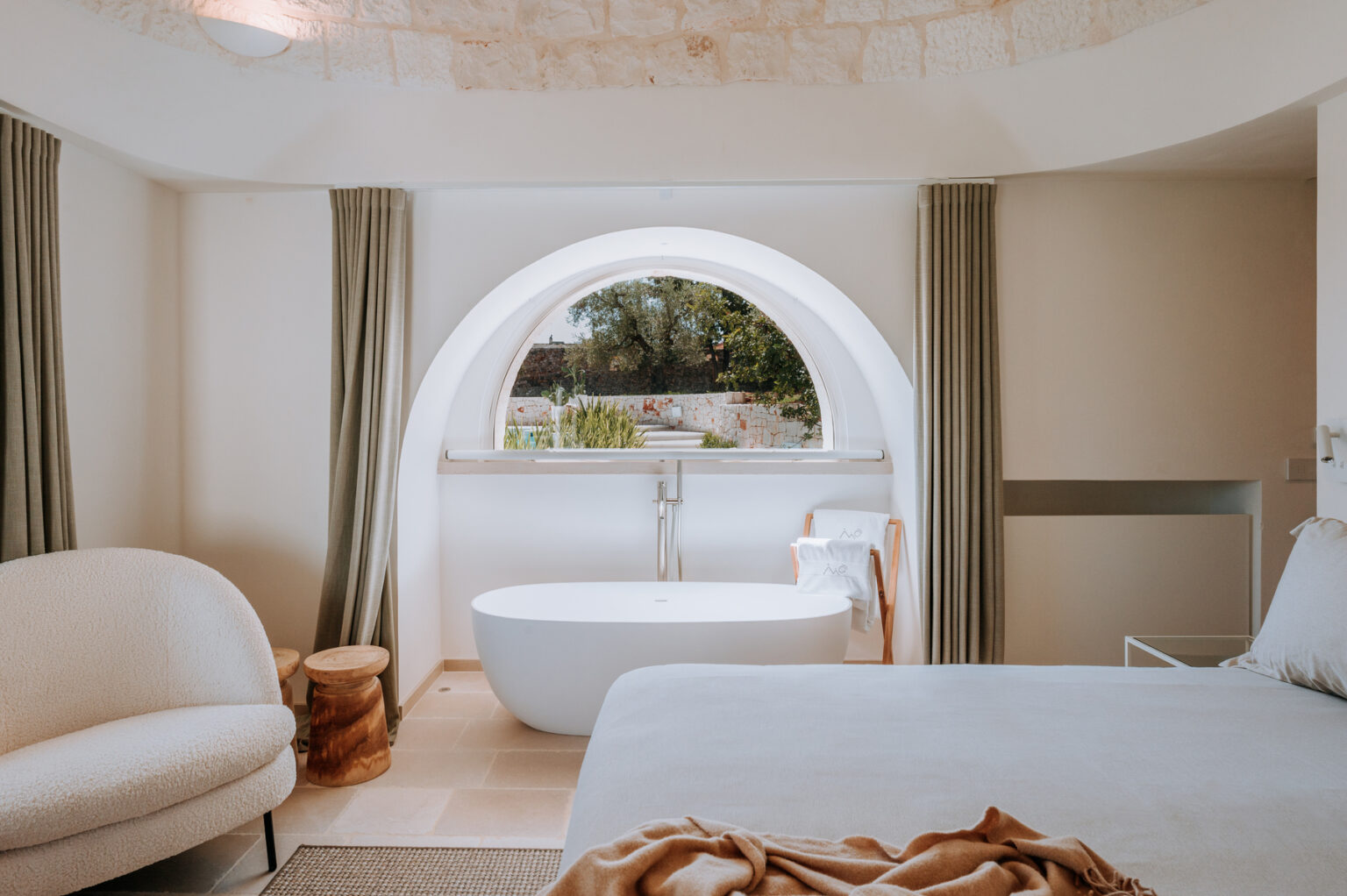 Modern bedroom interior featuring a cozy seating area, a freestanding bathtub, and large circular window offering a scenic view. The design includes neutral tones, stone accents, and soft textiles, creating a tranquil atmosphere. | Only on SopranoVillas
