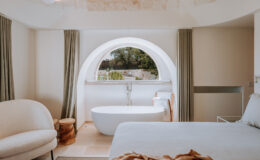 Modern bedroom interior featuring a cozy seating area, a freestanding bathtub, and large circular window offering a scenic view. The design includes neutral tones, stone accents, and soft textiles, creating a tranquil atmosphere.