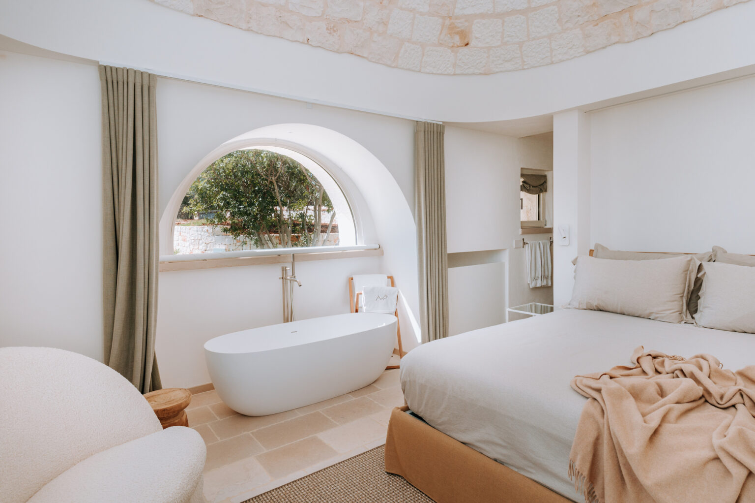 Modern and elegant bedroom featuring a freestanding bathtub, neutral-toned bedding, and large arched windows that offer natural light and a view of greenery. The minimalist decor includes soft curtains and a cozy armchair, creating a serene and inviting atmosphere. | Only on SopranoVillas