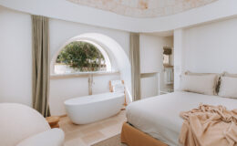 Modern and elegant bedroom featuring a freestanding bathtub, neutral-toned bedding, and large arched windows that offer natural light and a view of greenery. The minimalist decor includes soft curtains and a cozy armchair, creating a serene and inviting atmosphere.