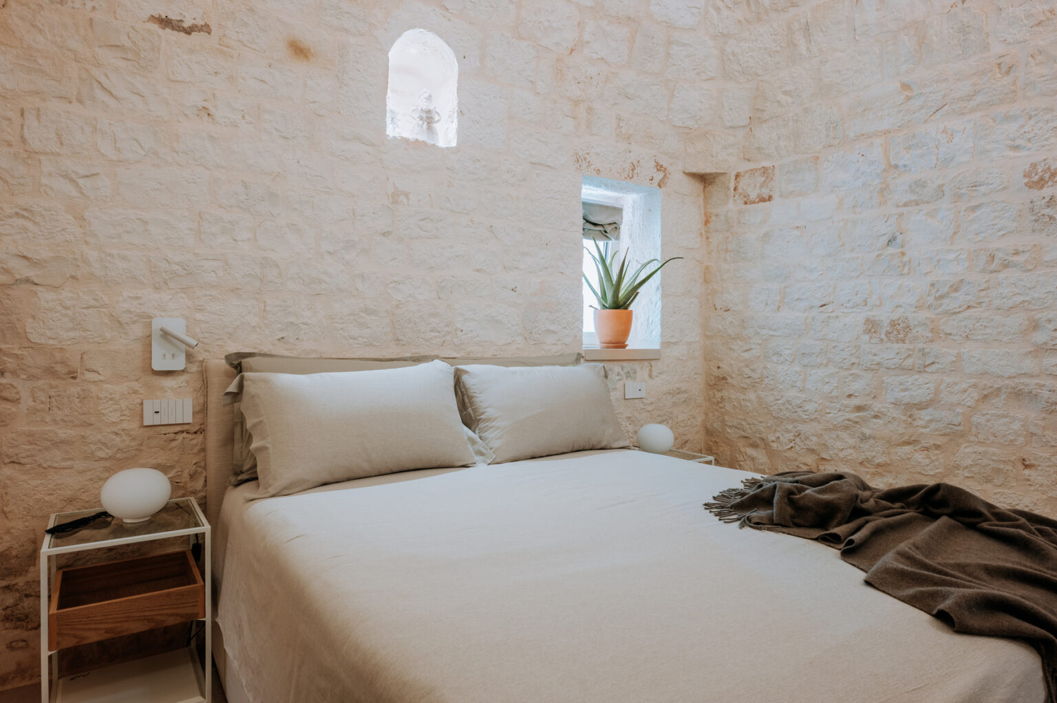 Cozy bedroom featuring a minimalist design with stone walls, a comfortable bed with neutral linens, a small bedside table, and a potted plant by the window. | Only on SopranoVillas