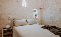 Cozy bedroom featuring a minimalist design with stone walls, a comfortable bed with neutral linens, a small bedside table, and a potted plant by the window.